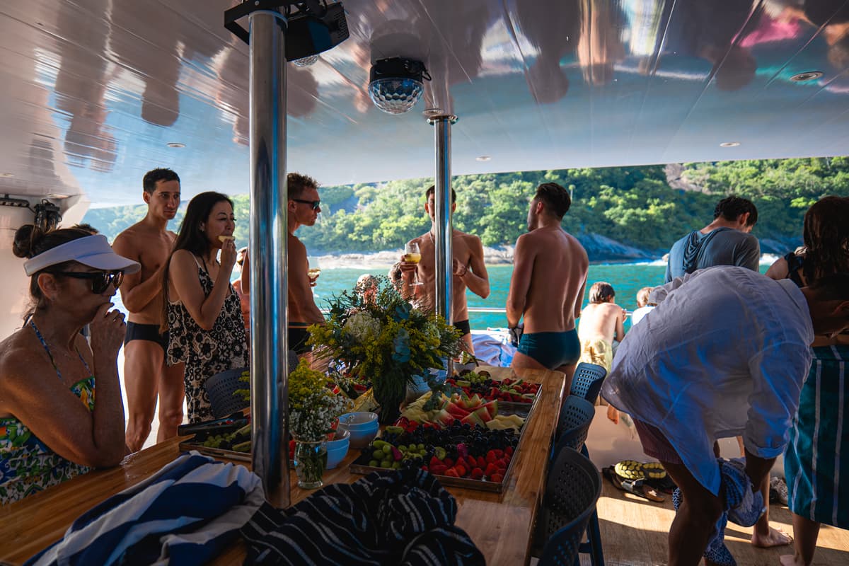 Convidados a bordo em torno de mesa de frutas com a costa verde ao fundo